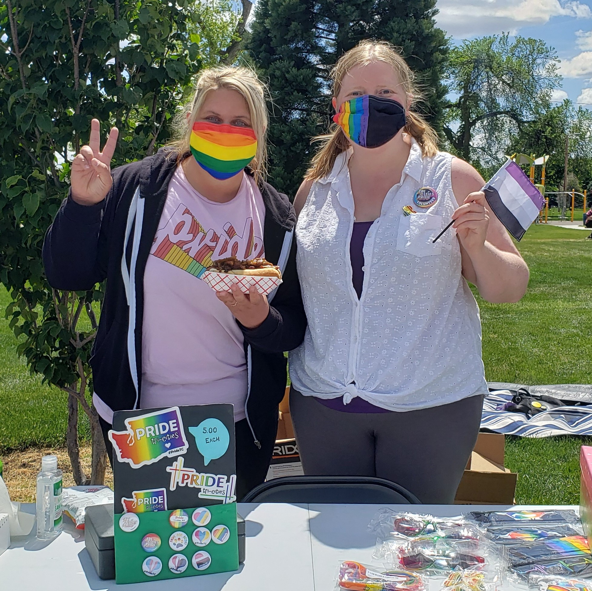 Kim and a friend are standing behind a table at a Pride event wearing rainbow masks on a sunny day.