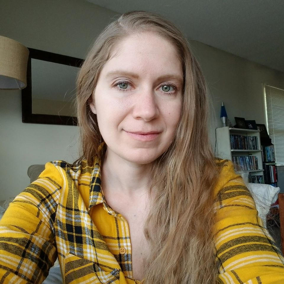 Kelly is a thirty something woman with light skin and long dark blonde hair. She is wearing a yellow and black plaid shirt and smiling.