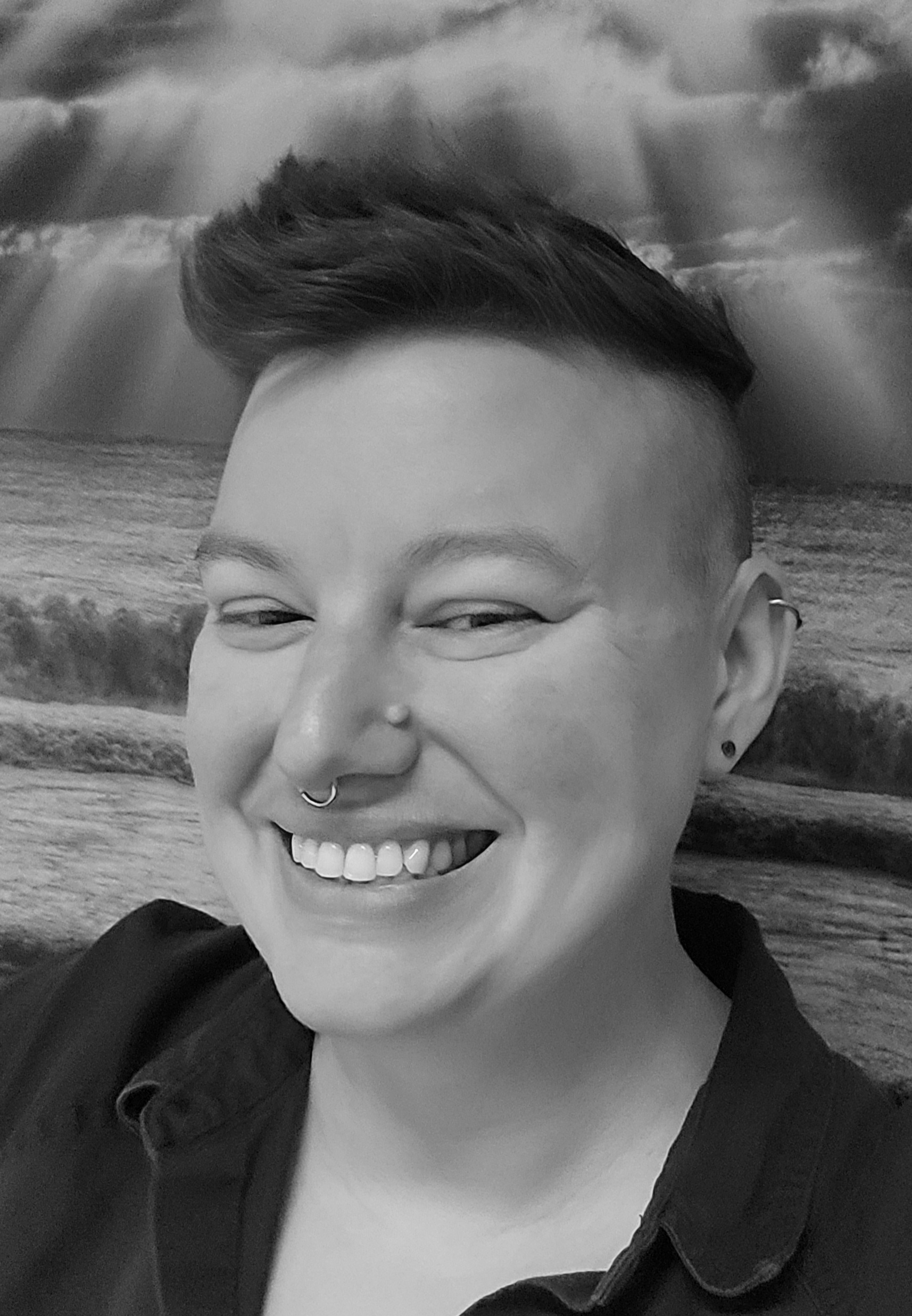 Heidi is a twenty something caucasian person with dark hair that is a few inches long on top and shaved on the sides. They are wearing a dark collared shirt and are leaning against a landscape picture of a beach scene while smiling. They have a septum piercing and two ear piercings.
