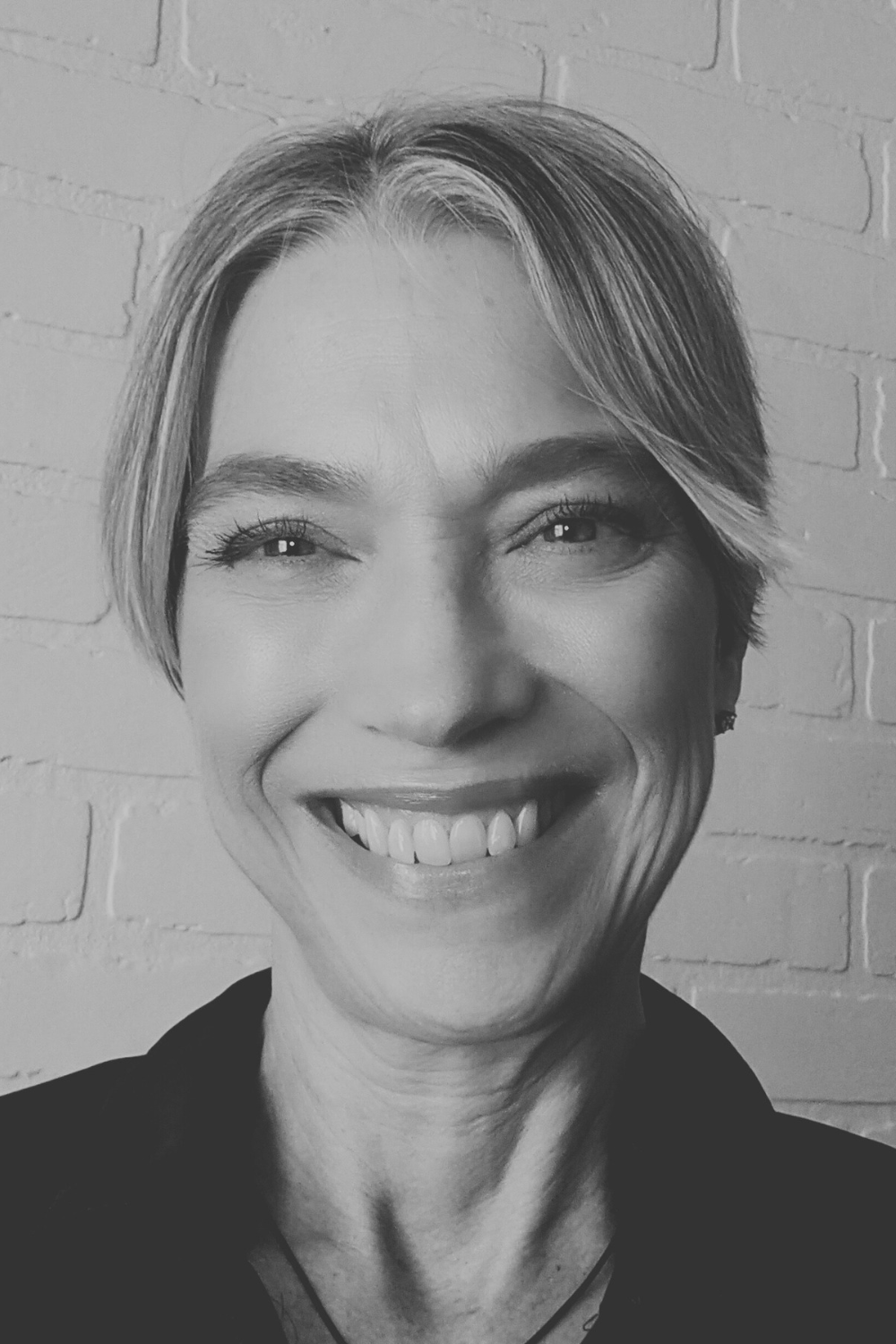 This black and white photo shows Keri, a middle-aged caucasian female with blonde hair that is pulled back behind her head and parted in the middle. She is wearing a dark-colored sweater or jacket and is smiling warmly.