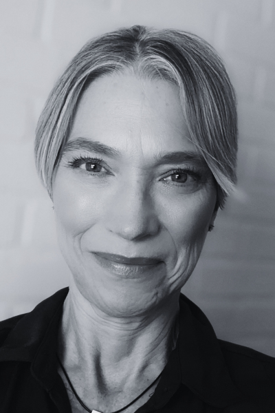 This black and white image features Keri, a middle-aged Caucasian woman with light colored hair parted in the middle and pulled back behind her head. She is smiling warmly with a closed mouth and wearing a black collared top.