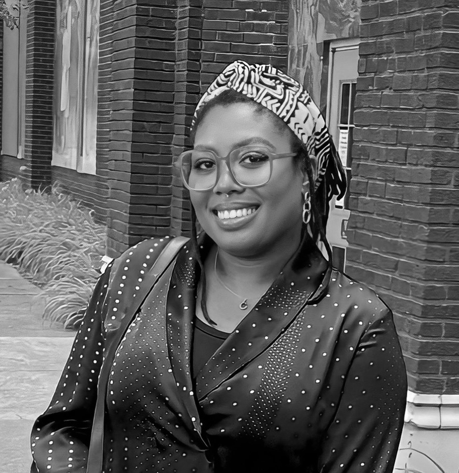 Briana is a twenty something African American woman. She is pictured outside a brick building wearing a headscarf, glasses, and a dark colored blazer with small polka dots, and a purse slung over her right shoulder. Her body is positioned facing just to the viewer's left and she is looking at the camera and smiling.