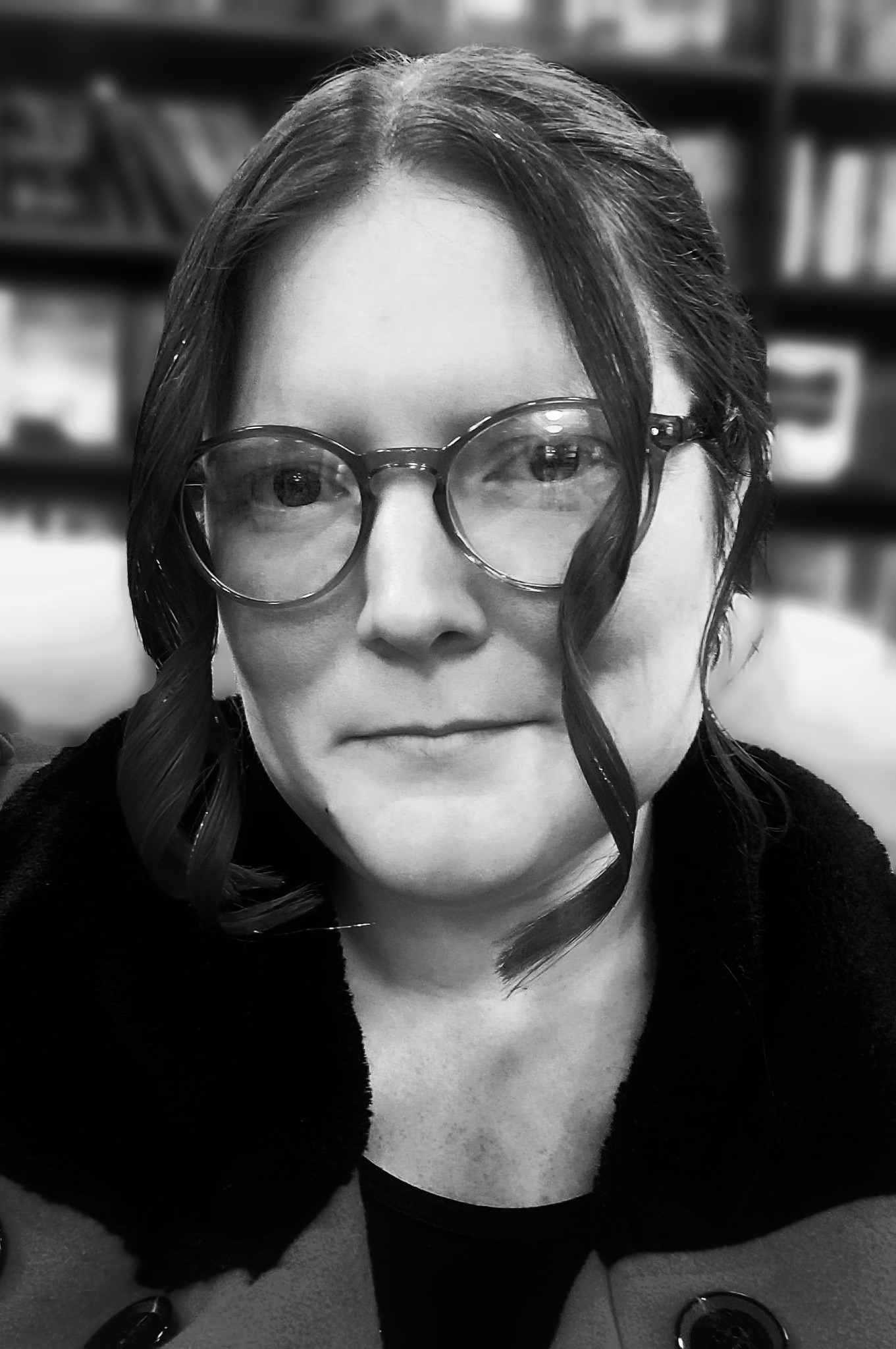 This black and white photo features Kim, a thirty-something year old Caucasian woman. She is wearing glasses, a coat with a dark, wide collar, and has her dark hair pulled back with two pieces hanging loose on either side of her face that are slightly curled. Her head is tilted slightly to the viewer's left and she has a neutral expression.