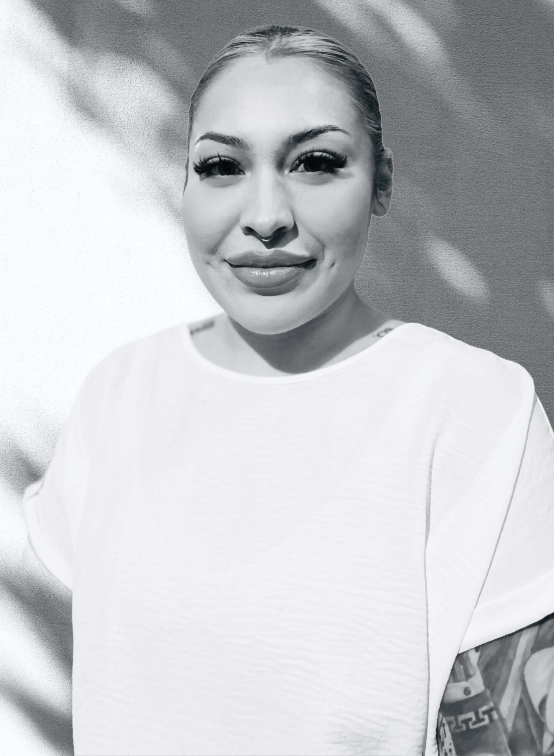 This black and white photo features Paola, a twenty something Hispanic woman. She is wearing a white t-shirt, has her bleached hair pulled up into a bun hidden behind her head and has a visible sleeve tattoo on her left arm. She has long, full lashes, full lips and dimpled cheeks. She is standing at a slight angle and looking at the camera with a closed mouth smile. She is in front of a while textured background with dappled sunlight on it.