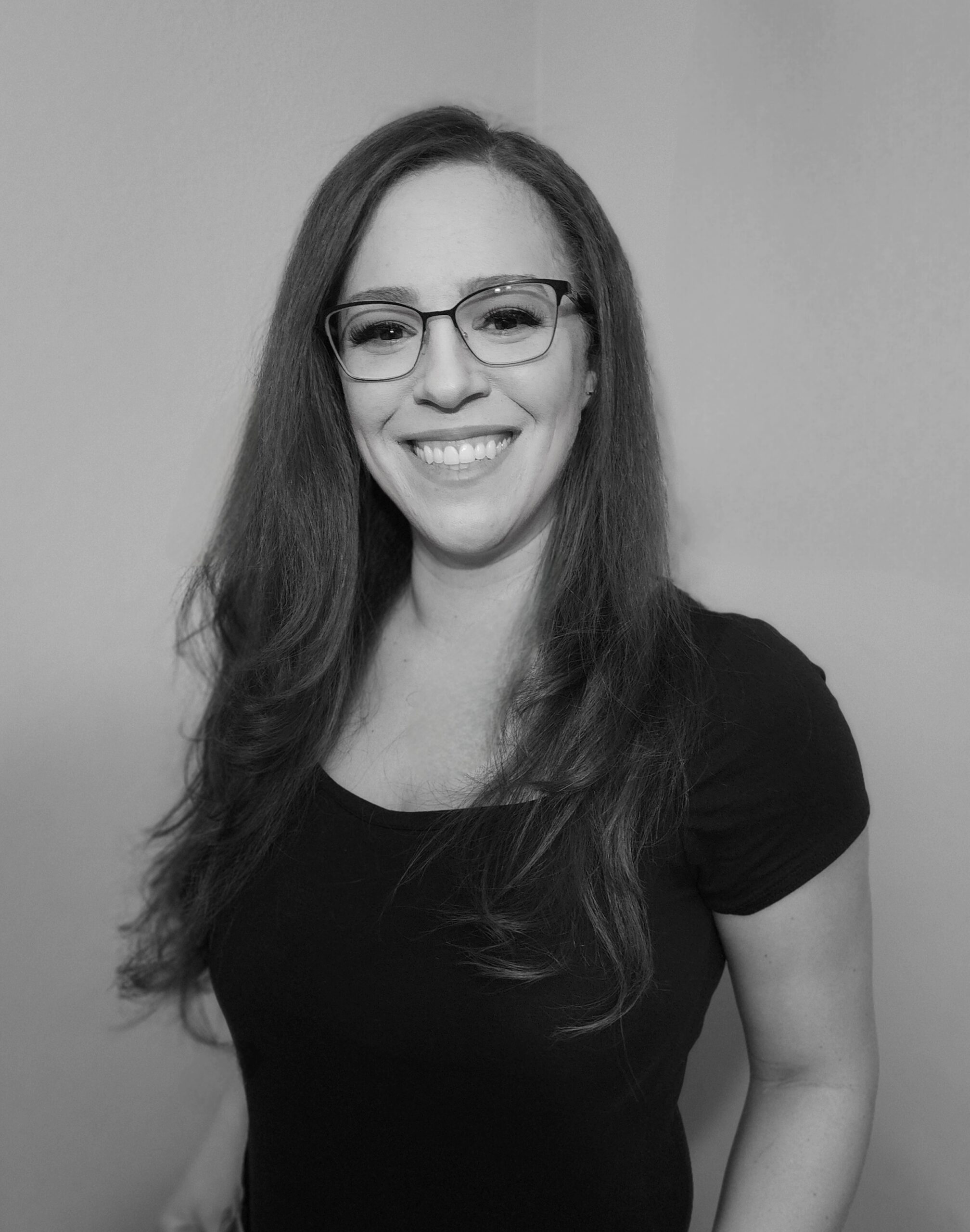 This black and white photo features a waist-up shot of Taneisha, a twenty something woman with long dark hair and glasses, who is wearing a dark-colored fitted t-shirt. She is standing at an angle and looking straight ahead at the viewer and smiling warmly with an open mouth. She is standing against a neutral-colored background.