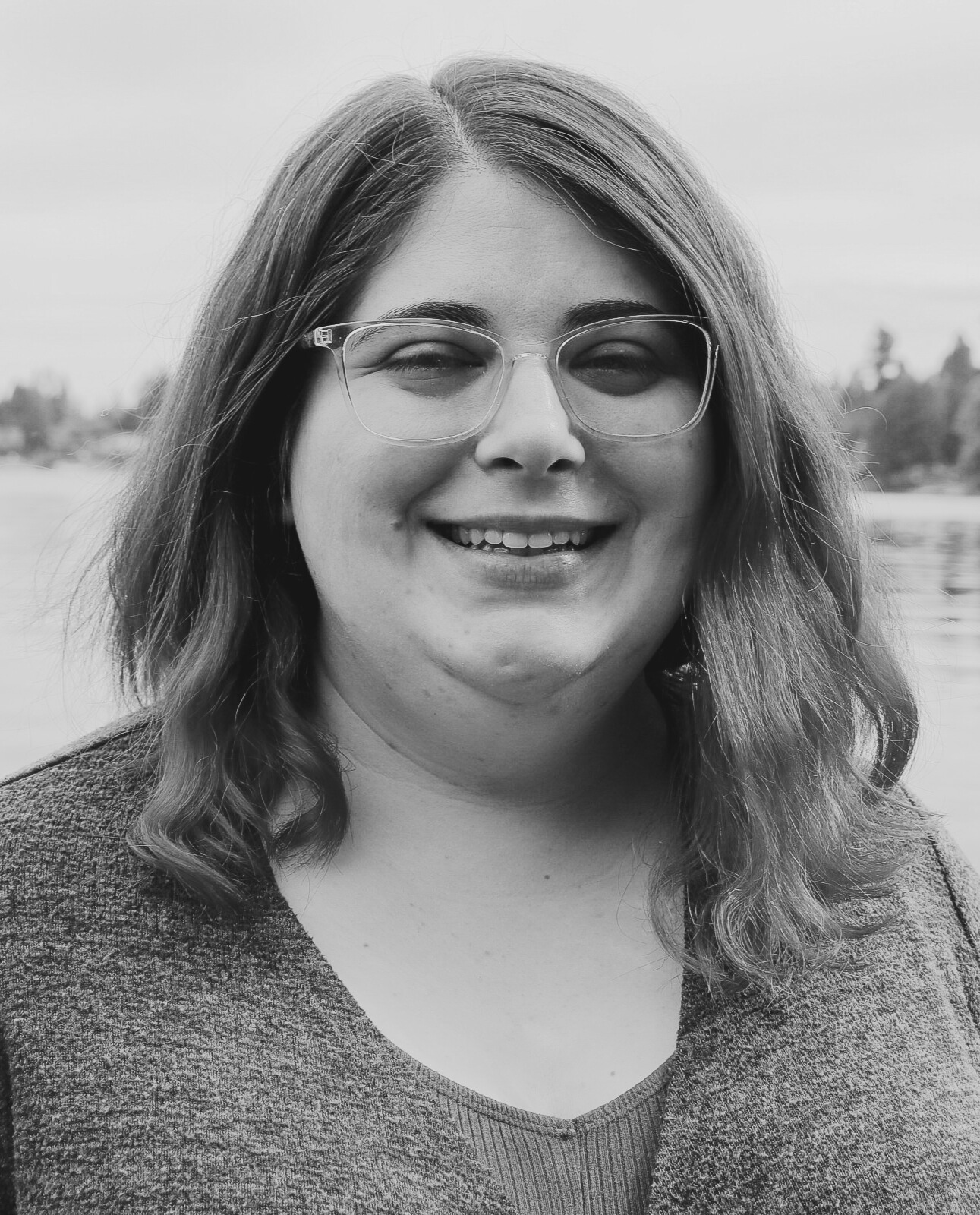 This black and white photo features Kaylynn, a twenty something year old heavy-set female with medium-colored hair just past her shoulders that curls at the ends. She is wearing glasses and a medium-colored sweater and is smiling warmly with an open mouth. She is pictured outside in front of a river on an overcast day.