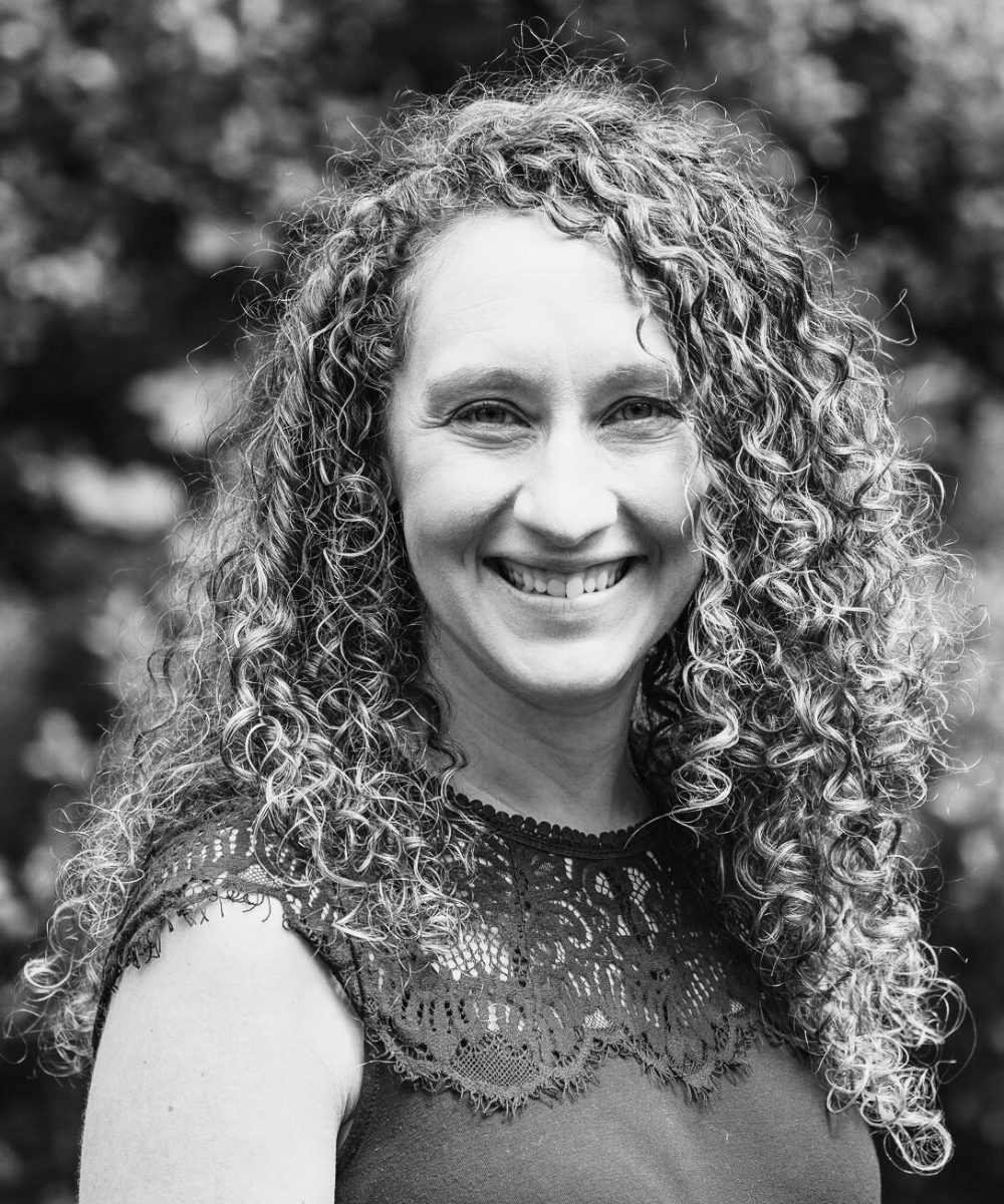 This image shows Nancy, a 40 something year old woman with curly light-colored hair that descends past her shoulders. Her body is angled to the viewer's right and she is wearing a medium tone sleeveless shirt with the top portion being made of lace. She is looking straight at the camera and smiling warmly.