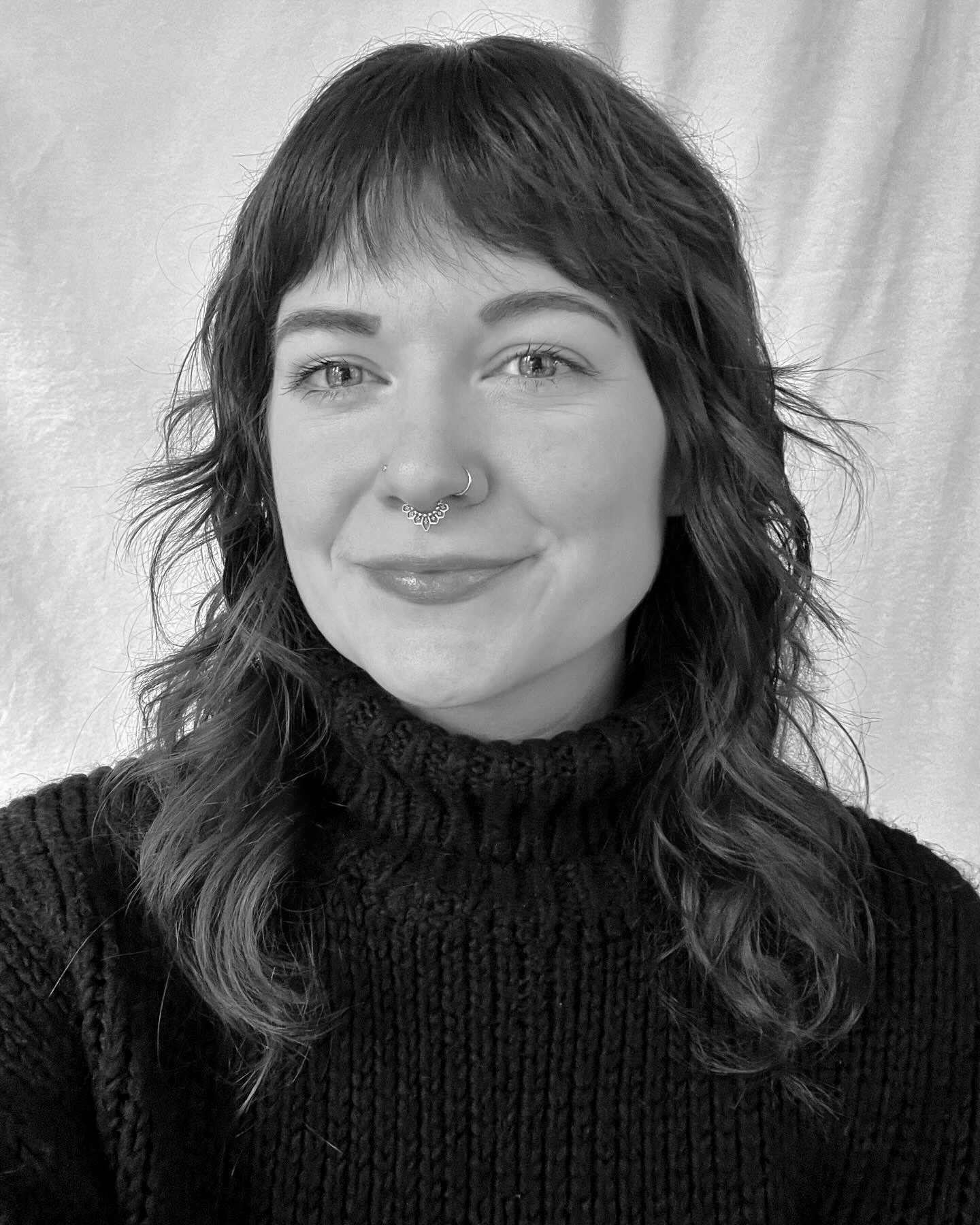 This black and white image features MacKinsey, a twenty something caucasian woman with medium-toned, shoulder-length, feathered hair with short bangs. She is wearing a dark colored turtleneck sweater and shown in front of a light-colored fabric background. Her face is turned slightly towards the viewer's left and she is smiling slightly with a closed mouth.