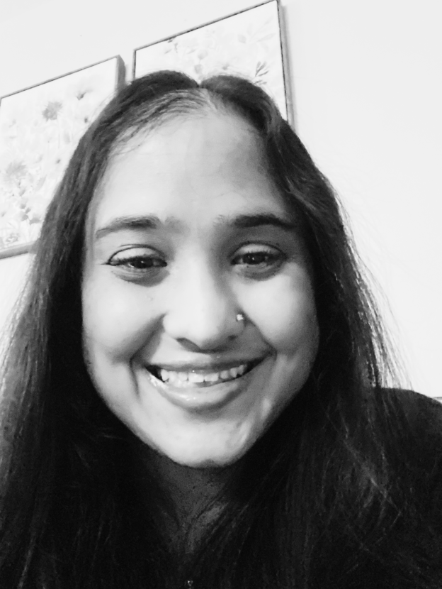 This black and white photo features Sapna, a twenty something, ethnic female with long dark hair, a nose ring, and an open mouth, warm smile. She is wearing a dark-colored shirt and is shown against a light-colored wall with art in the background.