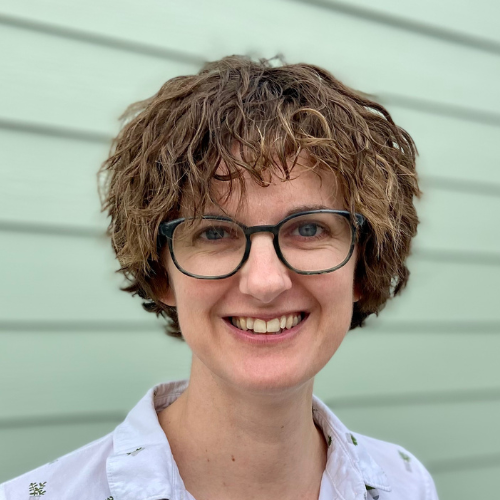 Hillary is a thirty-something caucasian woman with short curly brown hair. She is wearing glasses and a white button down shirt with a green plant pattern on it. She is looking at the camera and smiling warmly with an open mouth.