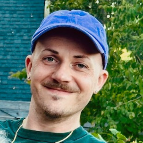 Vincent is a twenty-something, caucasian, male-presenting person. He has a brown moustache, a small patch of hair just below his bottom lip and a larger one on his chin. He is wearing a blue baseball cap, a green t-shirt, has small guages in his ears and is wearing a thin gold chain. He is pictured outside with green leaves behind him.