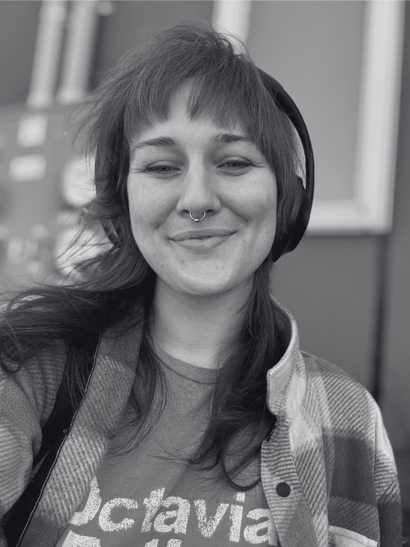 This image features Dee, a twenty something female-presenting queer person. Dee is smiling with a closed mouth and their eyes are open just a tiny bit. They are wearing a plaid shirt over a t-shirt and over-the-ears-style headphones. They are caucasian with shoulder lenth hair and bangs as well as a septum piercing.