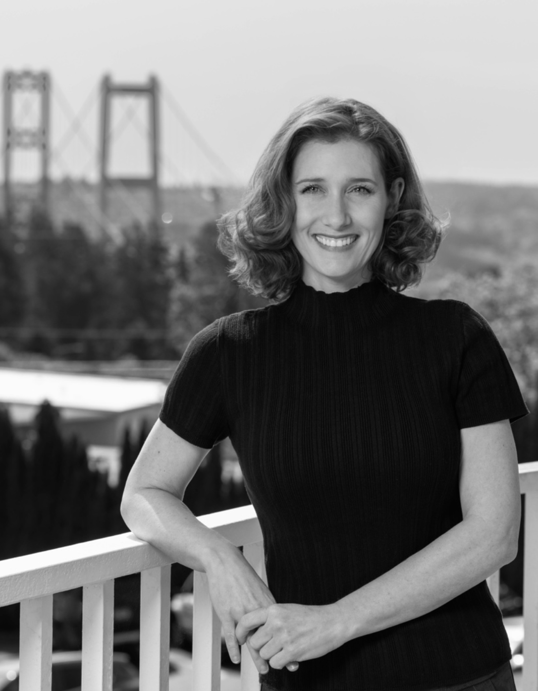 Leslie This iamge features Leslie, a slim, thirty something caucasian woman with medium length hair that is straight on the top, curls at the ends, and is parted over her left eye. She is wearing a short sleeve, dark-colored mock neck shirt and is leaning against a white fence on a sunny day with a bridge and river in a desert landscape background. She is smiling warmly with an open mouth.