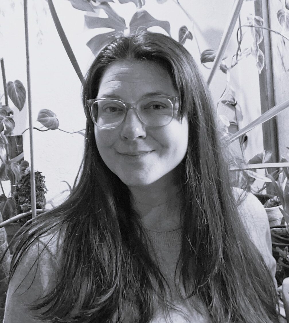 Lindsey is featured in this black and white photo. She is a caucasian woman appearing to be in her early to mid-thirties with long, straight, dark hair that is layered. She is wearing glasses and a long sleeved, light-colored shirt. She is seen sitting near a sunny window with lots of plants and a mural of a tree behind her. She is looking straight at the camera with her face turned slightly to her right and smiling subtly with a closed mouth.
