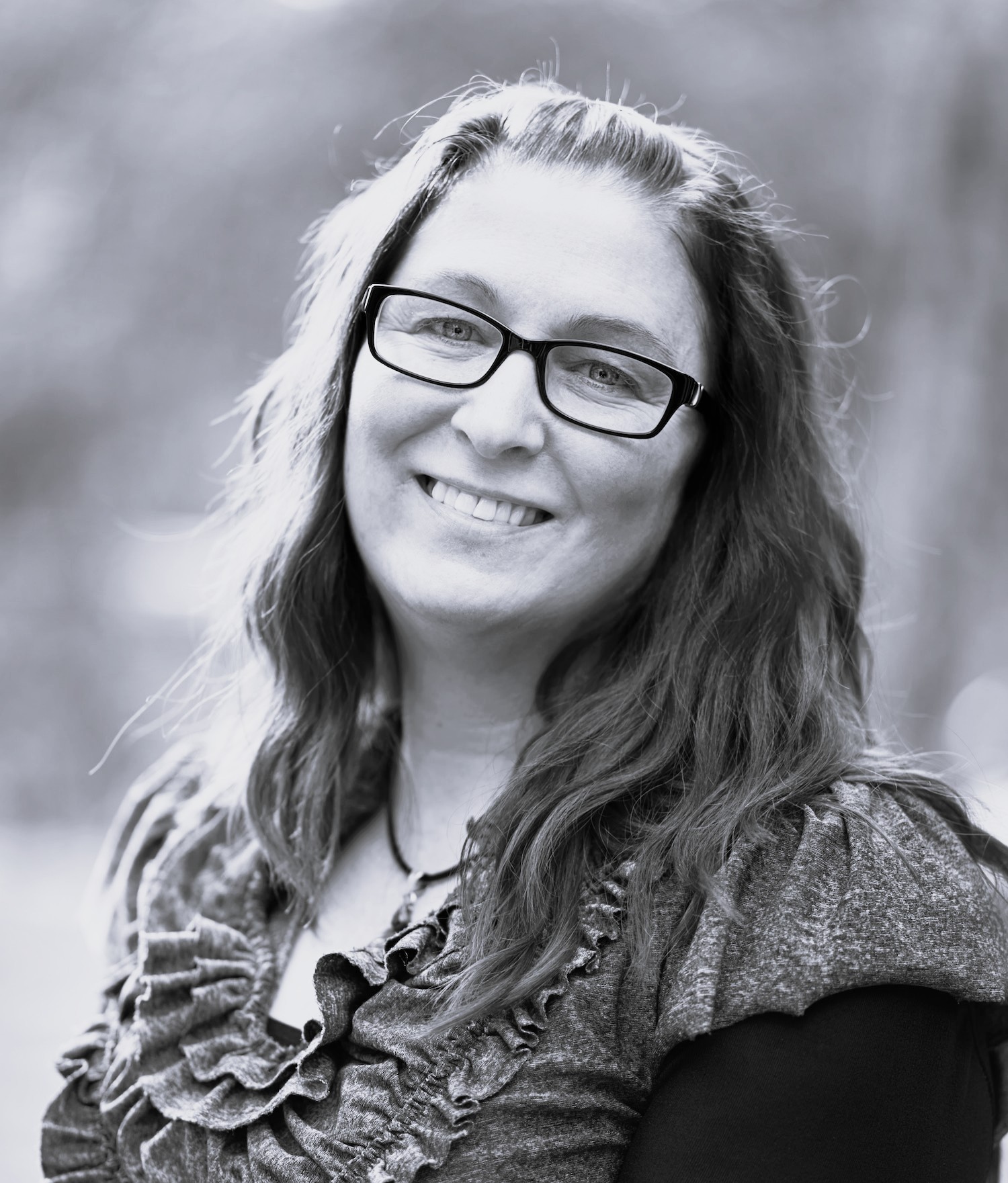 This black and white photo features Nicole, a late twenties to early thirties-aged caucasian woman. Nicole has long hair which softly curls, She is wearing a shirt with frilly trim around the neckline and black-rimmed glasses. She is positioned at an angle from the viewer. Her head is tilted toward the viewer's right and she is smiling with an open mouth.